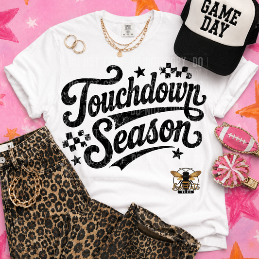 A styled flat-lay of a white graphic T-shirt with the words 'Touchdown Season' in large black script, accessorized with leopard-print shorts, a black-and-white cap reading 'GAME DAY,' gold rings, layered gold necklaces, a pink football, and a pink pom-pom on a pink star-patterned background.