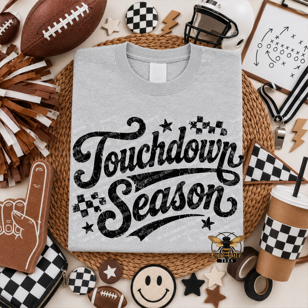 A light gray crewneck t-shirt with a black distressed 'Touchdown Season' graphic, presented in a flat-lay arrangement of football-themed props including a football, cheerleader pom-poms, a foam finger, and checkered accessories.