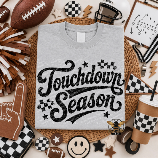 A light gray crewneck t-shirt with a black distressed 'Touchdown Season' graphic, presented in a flat-lay arrangement of football-themed props including a football, cheerleader pom-poms, a foam finger, and checkered accessories.