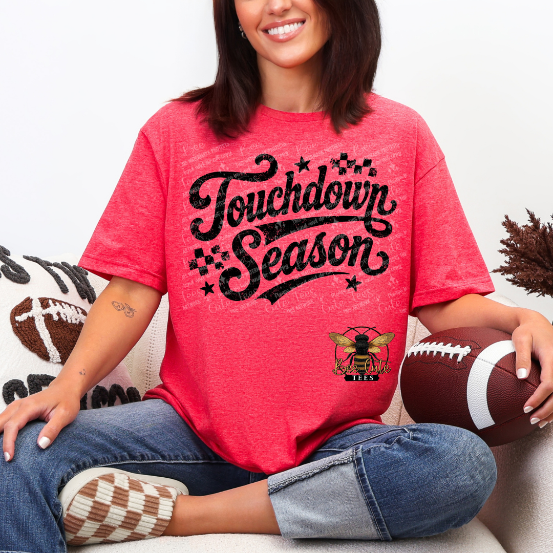A woman sitting on a couch wearing a  heathered red t-shirt with a black distressed 'Touchdown Season' graphic and a bee logo. She is holding a football.