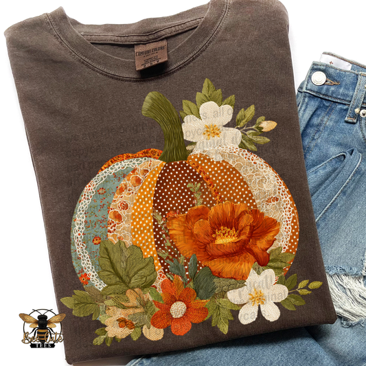 Folded vintage brown Comfort Colors t-shirt featuring a cottage-style pumpkin design with faux patchwork fabrics, delicate lace accents, and embroidered floral details. Warm shades of burnt orange, rust, cream, teal, and olive green create a cozy fall look. Styled with distressed blue jeans and a Bee Cute Tees watermark.