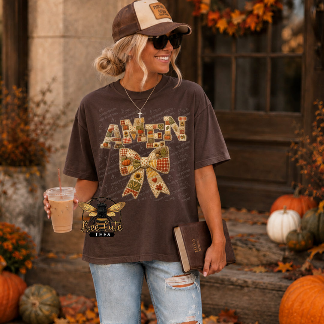 A woman stands outdoors wearing a brown graphic T-shirt with a patchwork-style 'AMEN' bow design, light blue ripped jeans, a tan cap, sunglasses, and holding an iced coffee and a book. Pumpkins and autumn decor are visible in the background.