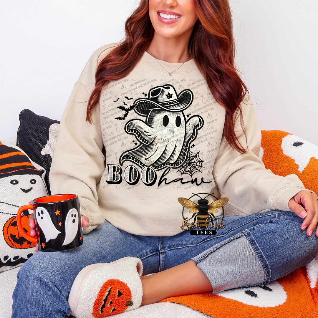 A woman wearing a beige crewneck sweatshirt with a black and white ghost graphic that says 'BOOHaw' sits on a cozy orange and white blanket. She is holding a black mug with a ghost design and wearing white slippers with orange pumpkin details.