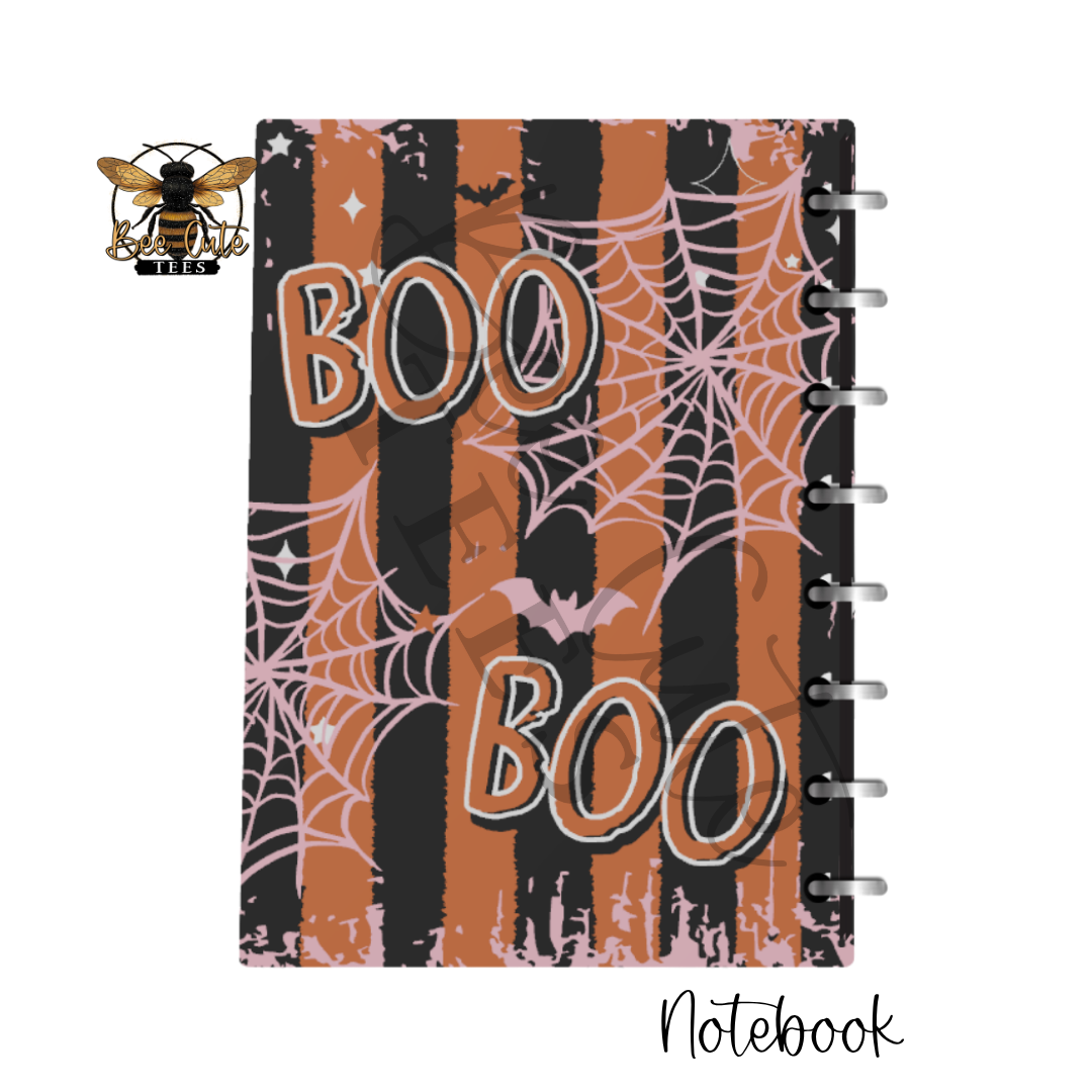 A spiral-bound notebook with a Halloween-themed cover featuring orange and black vertical stripes, pink spiderwebs, bats, and the words 'BOO BOO' in large outlined letters. A bee logo with 'BEE CUTE TEES' appears in the top-left corner, and 'Notebook' is written in script at the bottom-right on a white background.