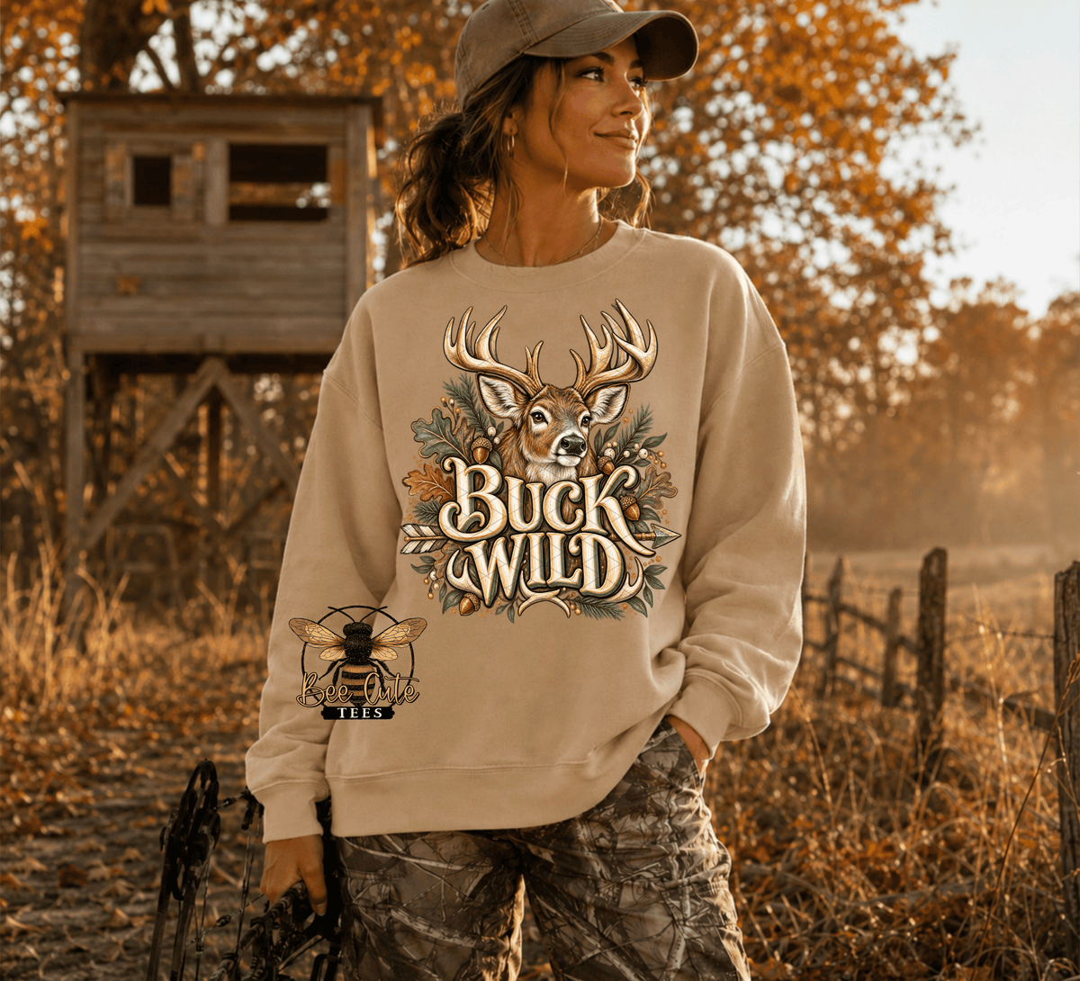 A woman wearing a beige crewneck sweatshirt with a 'Buck Wild' deer graphic and a bee patch on the sleeve, standing in an outdoor autumn field with a wooden hunting stand in the background.