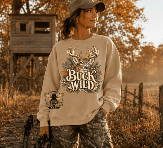 A woman wearing a beige crewneck sweatshirt with a 'Buck Wild' deer graphic and a bee patch on the sleeve, standing in an outdoor autumn field with a wooden hunting stand in the background.