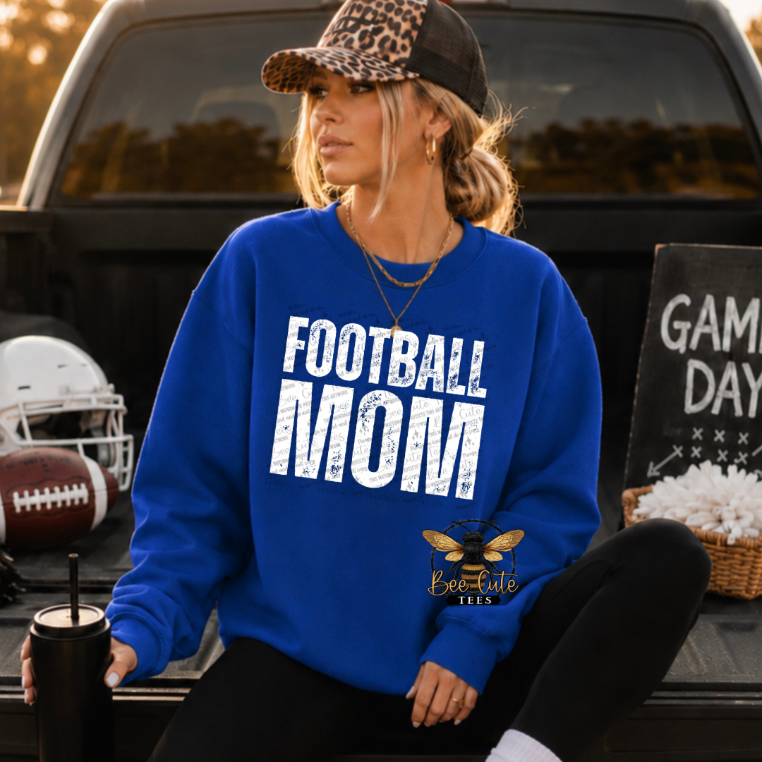 A woman sitting in the bed of a pickup truck wearing a royal blue crewneck sweatshirt with 'FOOTBALL MOM' printed in distressed white letters. She is wearing a leopard-print trucker hat and black pants.