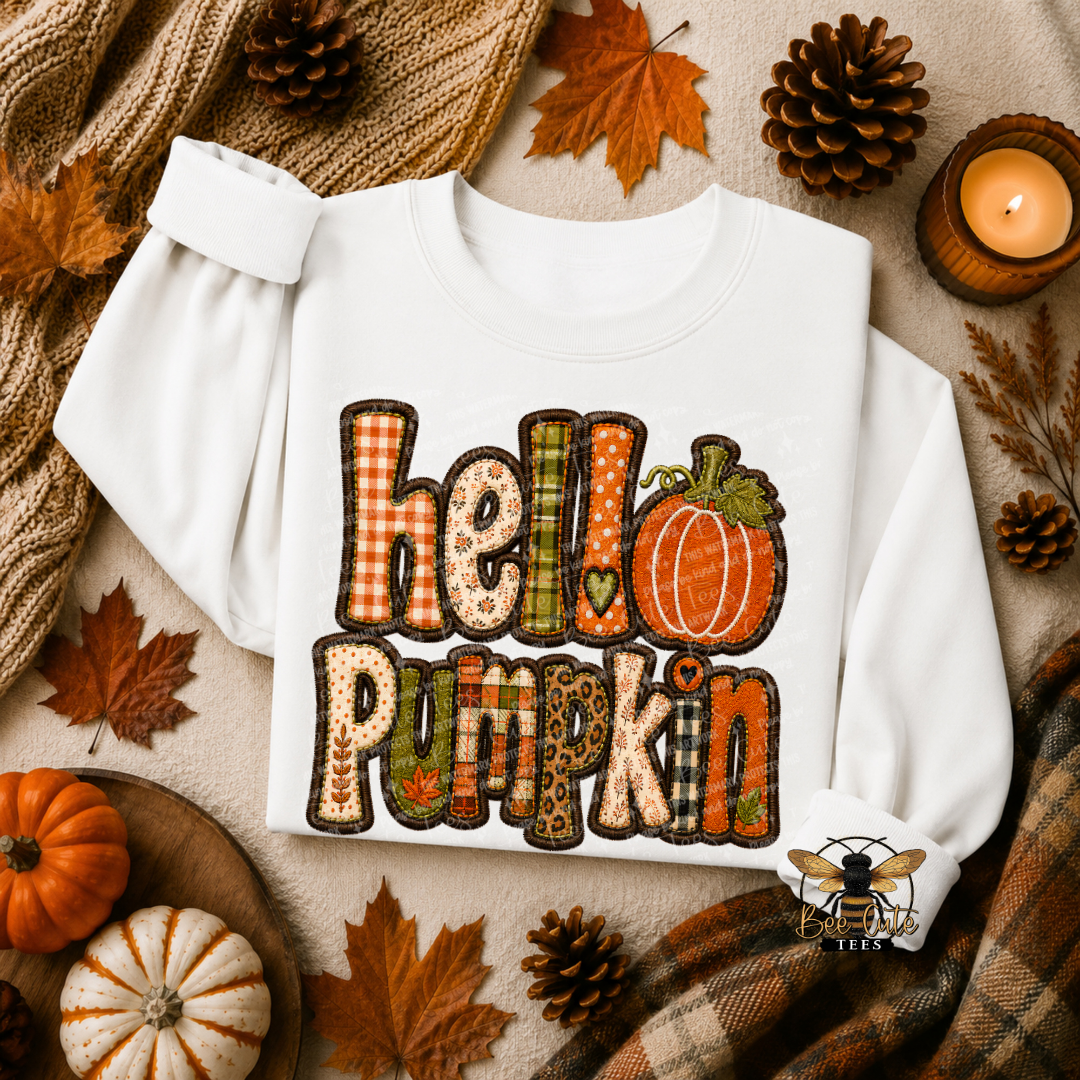 A white long-sleeve sweatshirt laid flat with a large autumn-themed 'hello pumpkin' graphic on the front. The graphic uses patchwork patterns in orange, brown, green, beige, and black.