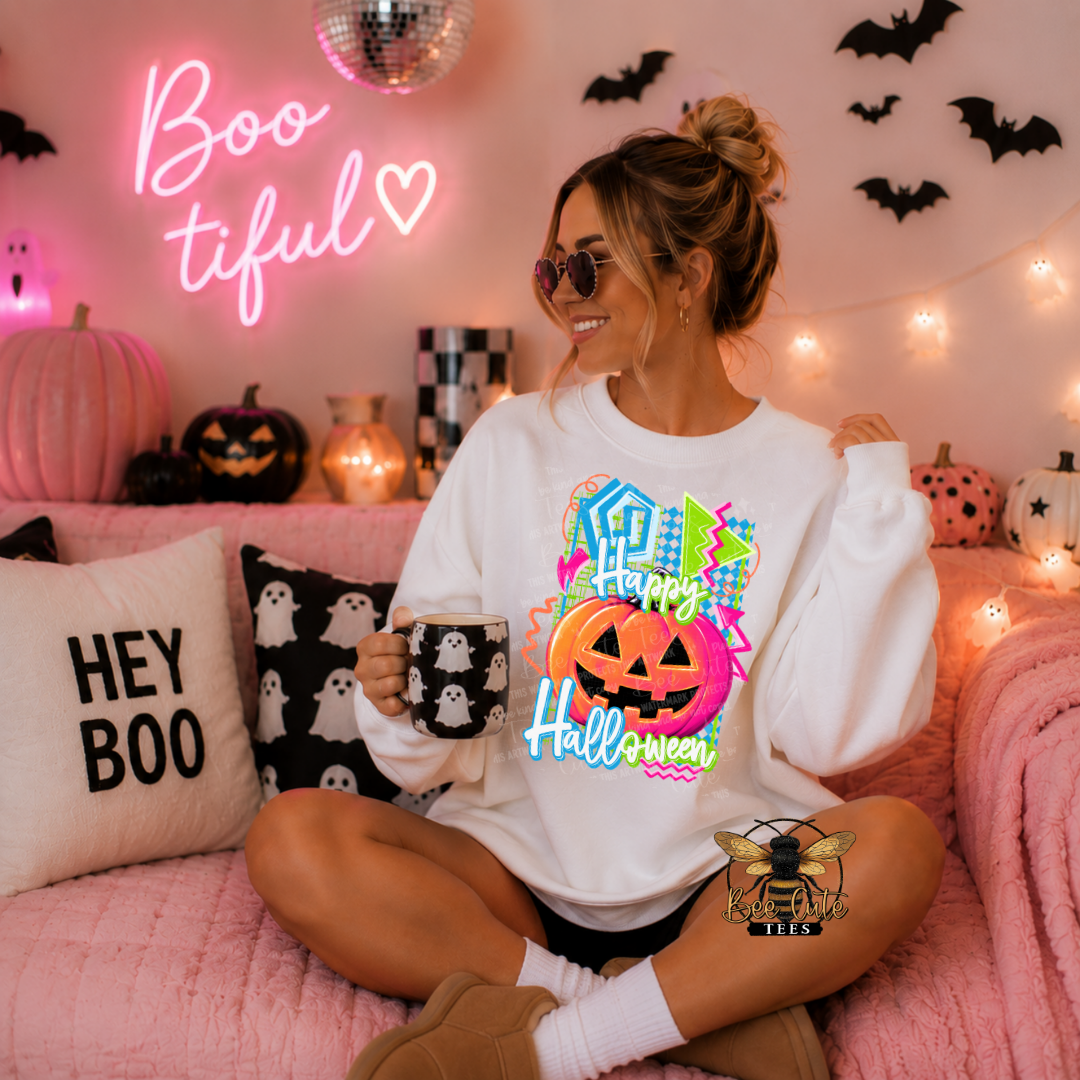 A woman wearing a white crewneck sweatshirt with a colorful 'Happy Halloween' graphic sits on a pink couch in a cozy, decorated Halloween room. The background features a pink neon sign, string lights, jack-o'-lanterns, and black bat decals.