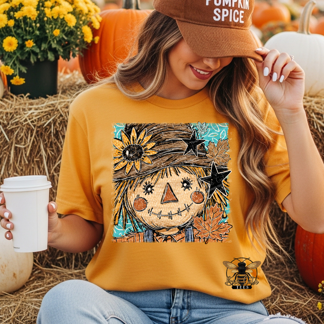 A woman wearing a mustard yellow graphic t-shirt with a scarecrow design, sitting in a pumpkin patch setting with hay bales and pumpkins.
