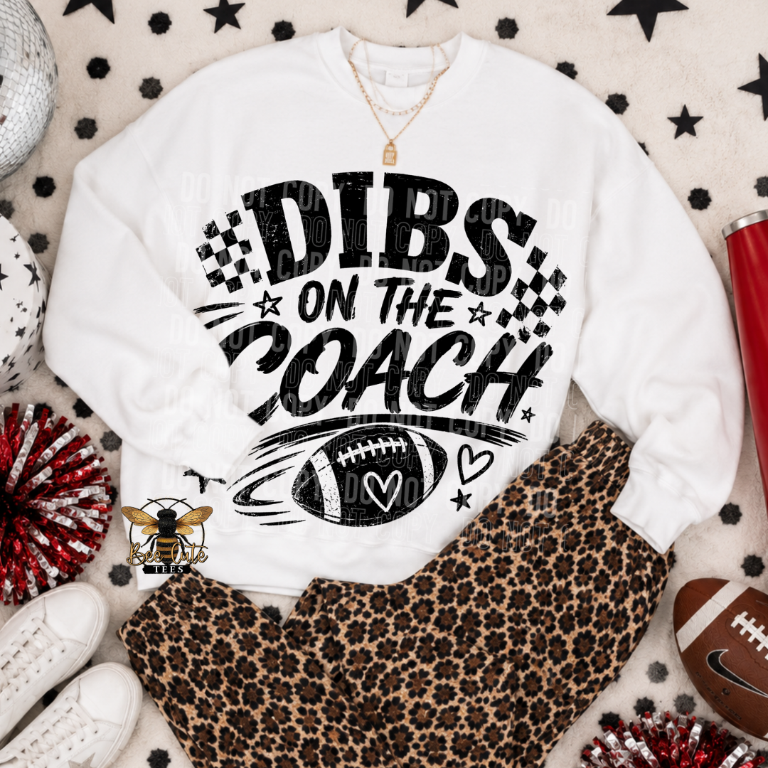 A white crewneck sweatshirt with a black graphic reading 'DIBS ON THE COACH' and a football design, styled in a flat lay with leopard print pants, white sneakers, a red tumbler, and decorative stars on a patterned blanket.