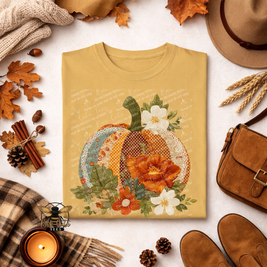 Folded mustard yellow t-shirt featuring a vintage patchwork pumpkin with floral fabrics, polka dots, faux embroidery textures, and delicate cream lace trim. Surrounded by orange and ivory flowers with olive green foliage. Styled with autumn leaves, a plaid scarf, candle, pinecones, and rustic accessories. Bee Cute Tees watermark visible.