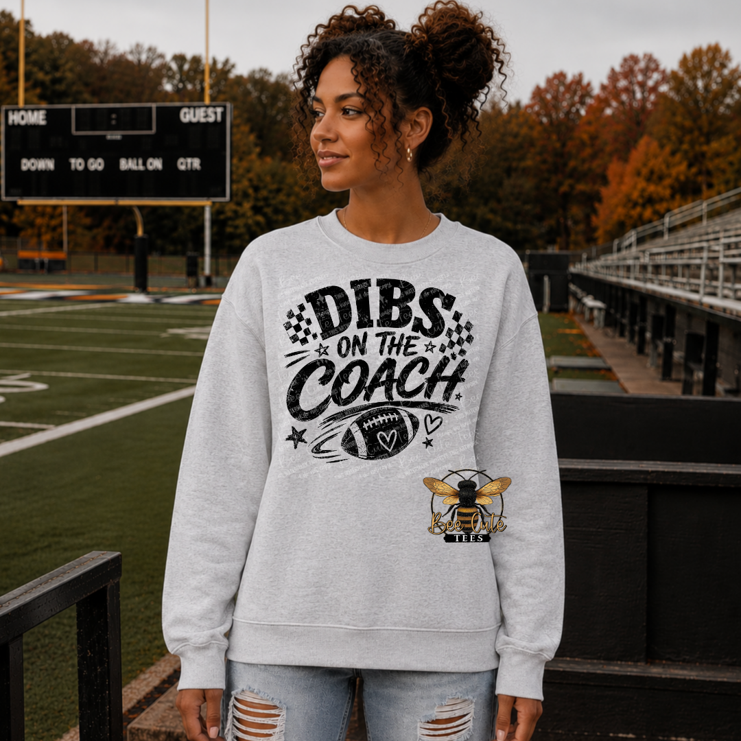A woman stands on a football field wearing a light gray crewneck sweatshirt with a black 'DIBS ON THE COACH' graphic and a bee logo on the lower right front. The background shows the green turf, stadium seating, and autumn trees.