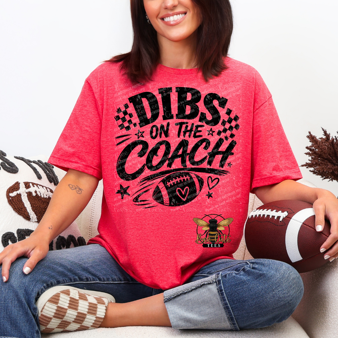 A woman sitting on a couch wearing a heather coral pink graphic t-shirt with 'DIBS ON THE COACH' and a football design in black. She is holding a brown football, with blue jeans and checkered socks visible.