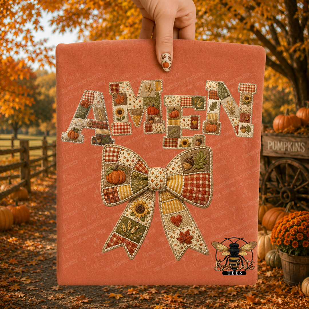 A hand holds up a peach-colored fabric item, likely a t-shirt or bandana, featuring a patchwork autumn-themed design with the word 'AMEN' in patchwork letters and a matching patchwork bow. The background is a blurred fall yard with pumpkins, orange foliage, and a wooden fence.