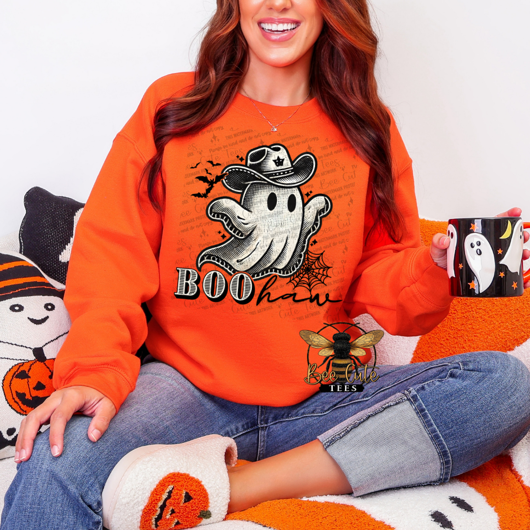 A woman wearing a bright orange crewneck sweatshirt with a Halloween graphic of a ghost in a cowboy hat and the word 'BOO haw'. She is sitting cross-legged on an orange and white blanket, holding a black mug with a white ghost design.