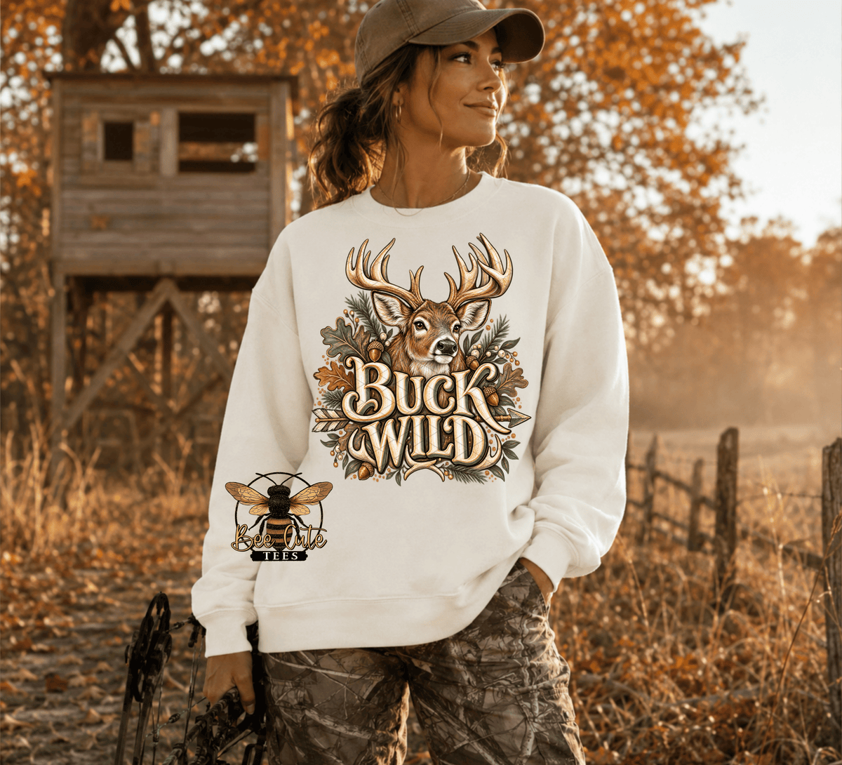 A woman wearing a white crewneck sweatshirt with a 'Buck Wild' deer graphic and a bee patch, standing in an outdoor autumn setting with a wooden hunting stand in the background.