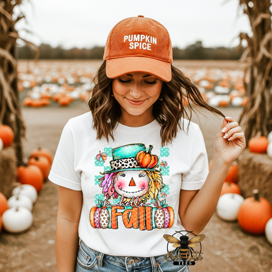 A woman standing in a pumpkin patch wearing a white graphic t-shirt with a colorful autumn scarecrow design and a rust-colored baseball cap that reads 'PUMPKIN SPICE'.