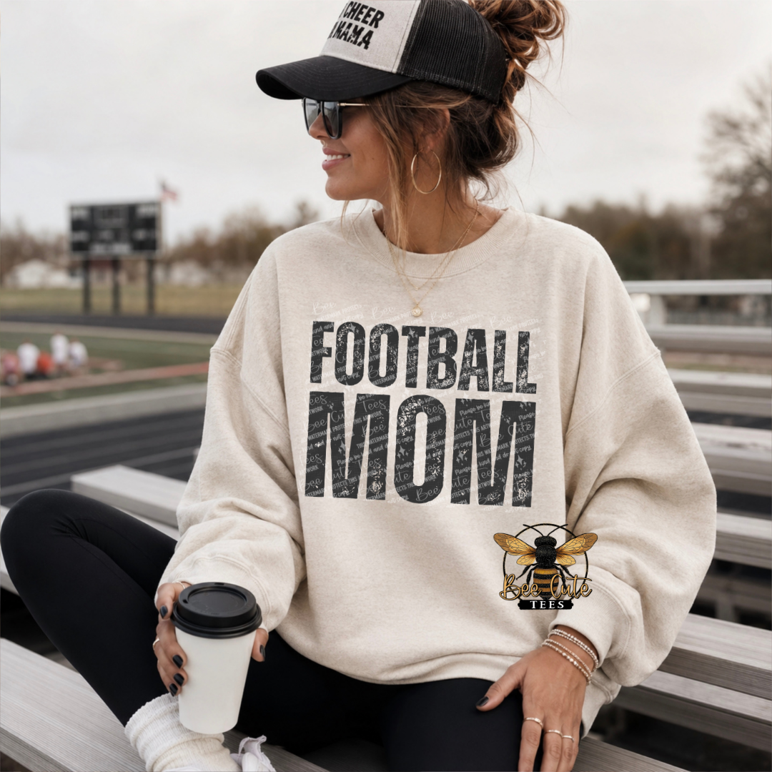 A woman sitting on outdoor bleachers wearing a cream sweatshirt with a large distressed black 'FOOTBALL MOM' graphic and a bee illustration, along with a black cap and sunglasses.