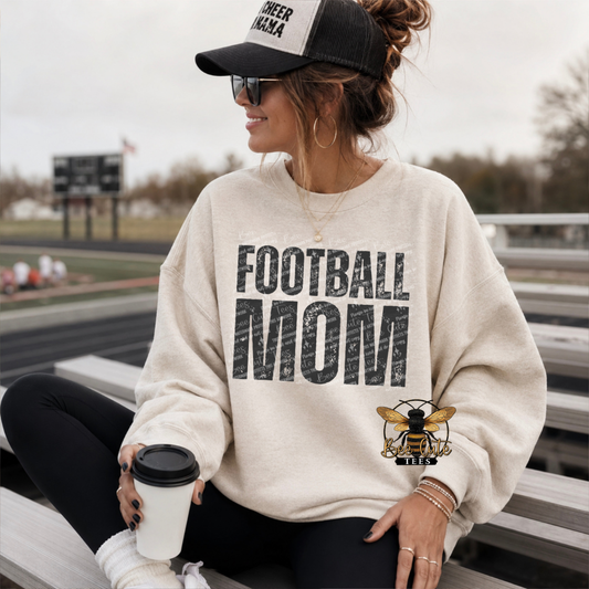 A woman sitting on outdoor bleachers wearing a cream sweatshirt with a large distressed black 'FOOTBALL MOM' graphic and a bee illustration, along with a black cap and sunglasses.