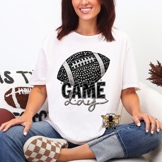 A woman wearing a white graphic t-shirt with a black and silver football design that reads 'GAME Day' and blue jeans, sitting on a couch.