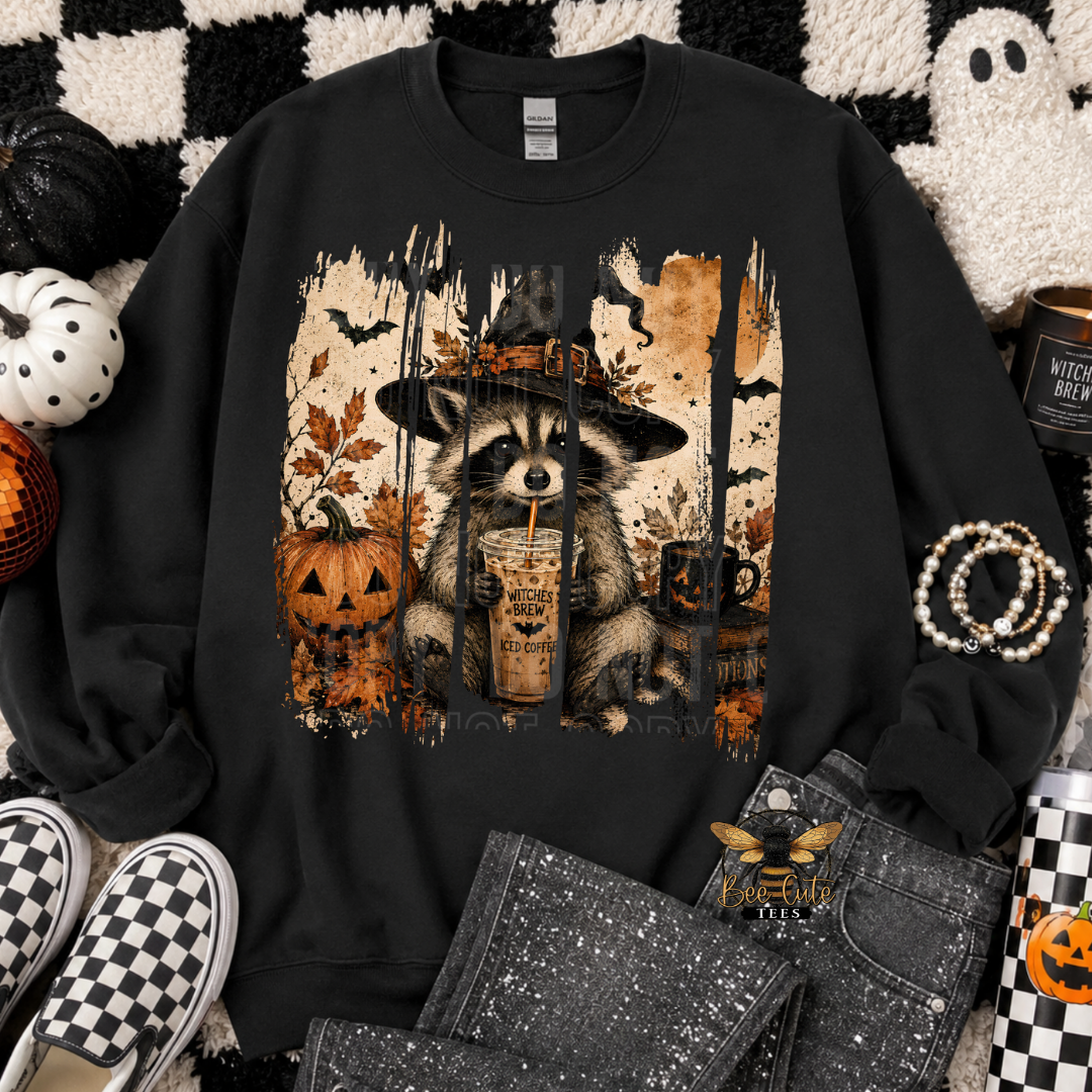 A black crewneck sweatshirt with a Halloween-themed graphic of a raccoon in a witch hat holding a pumpkin latte, styled in a flat-lay arrangement with jeans, slip-on shoes, and autumn props.
