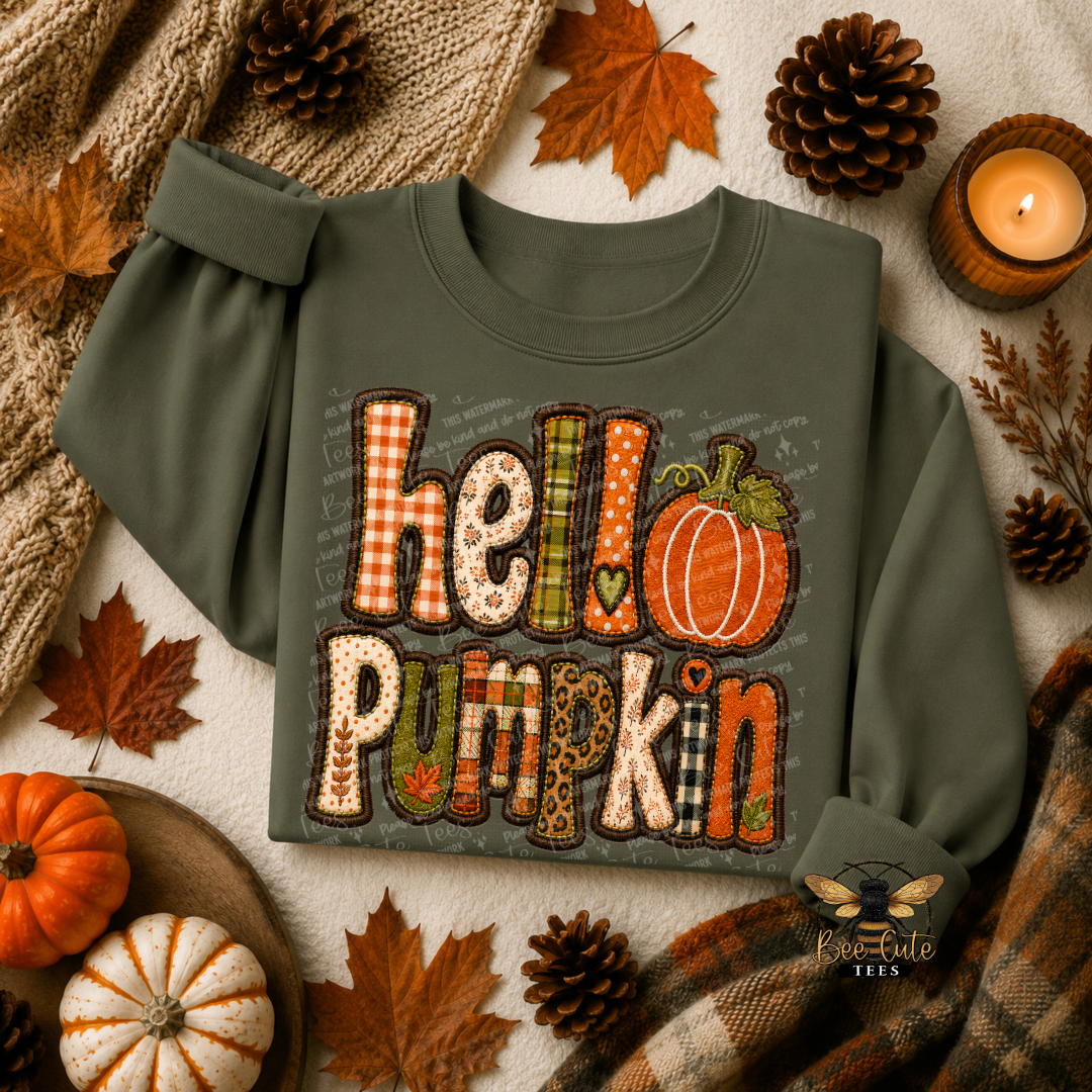 An olive green long-sleeve t-shirt with a 'hello pumpkin' graphic made of autumn-themed patterned letters, styled in a cozy fall flat-lay with pumpkins, pinecones, a candle, and a knit blanket.