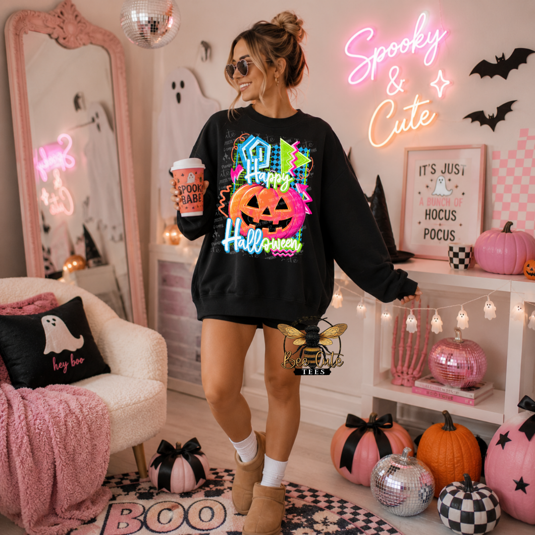 A woman stands in a decorated Halloween-themed room wearing a black crewneck sweatshirt with a colorful 'Happy Halloween' graphic featuring pumpkins and ghosts. She holds a coffee cup and wears shorts and tan boots, surrounded by pumpkins, a mirror, and neon signs.