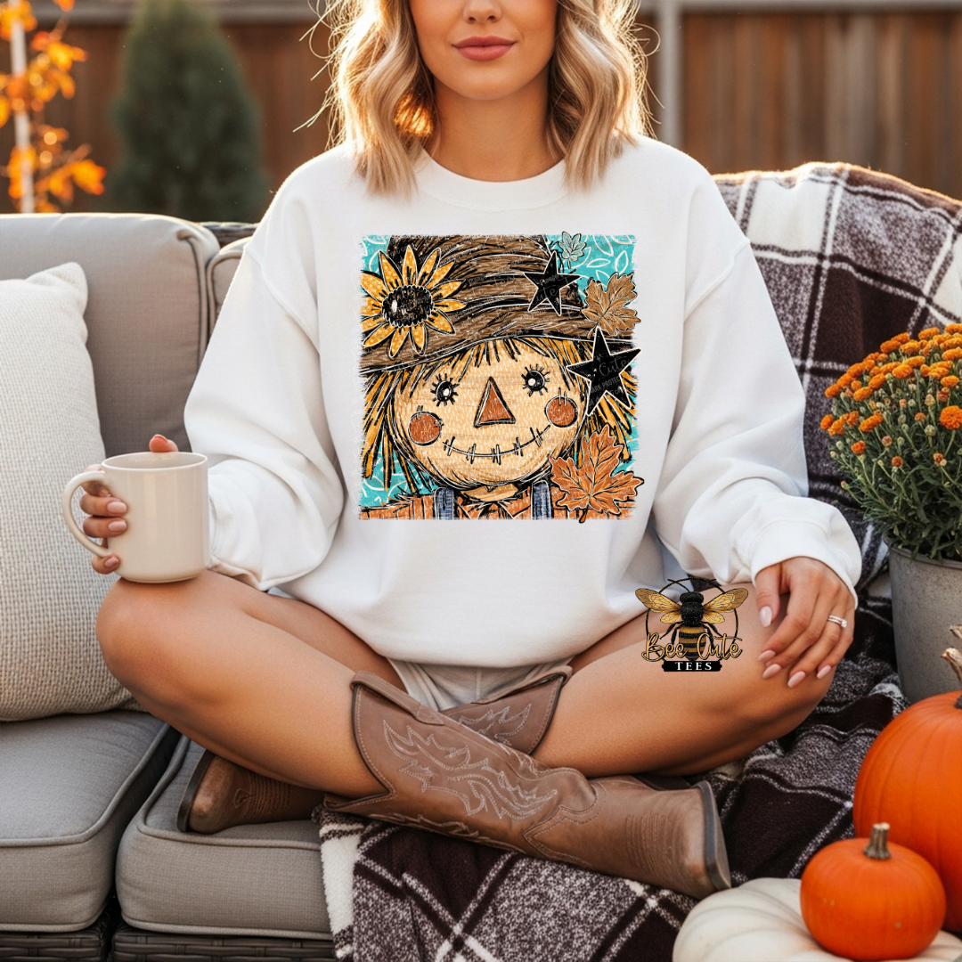 A woman sits cross-legged on a patio couch wearing a white crewneck sweatshirt with a colorful autumn scarecrow graphic on the front. She is holding a mug and wearing cowboy boots, with pumpkins and a plaid blanket in the background.