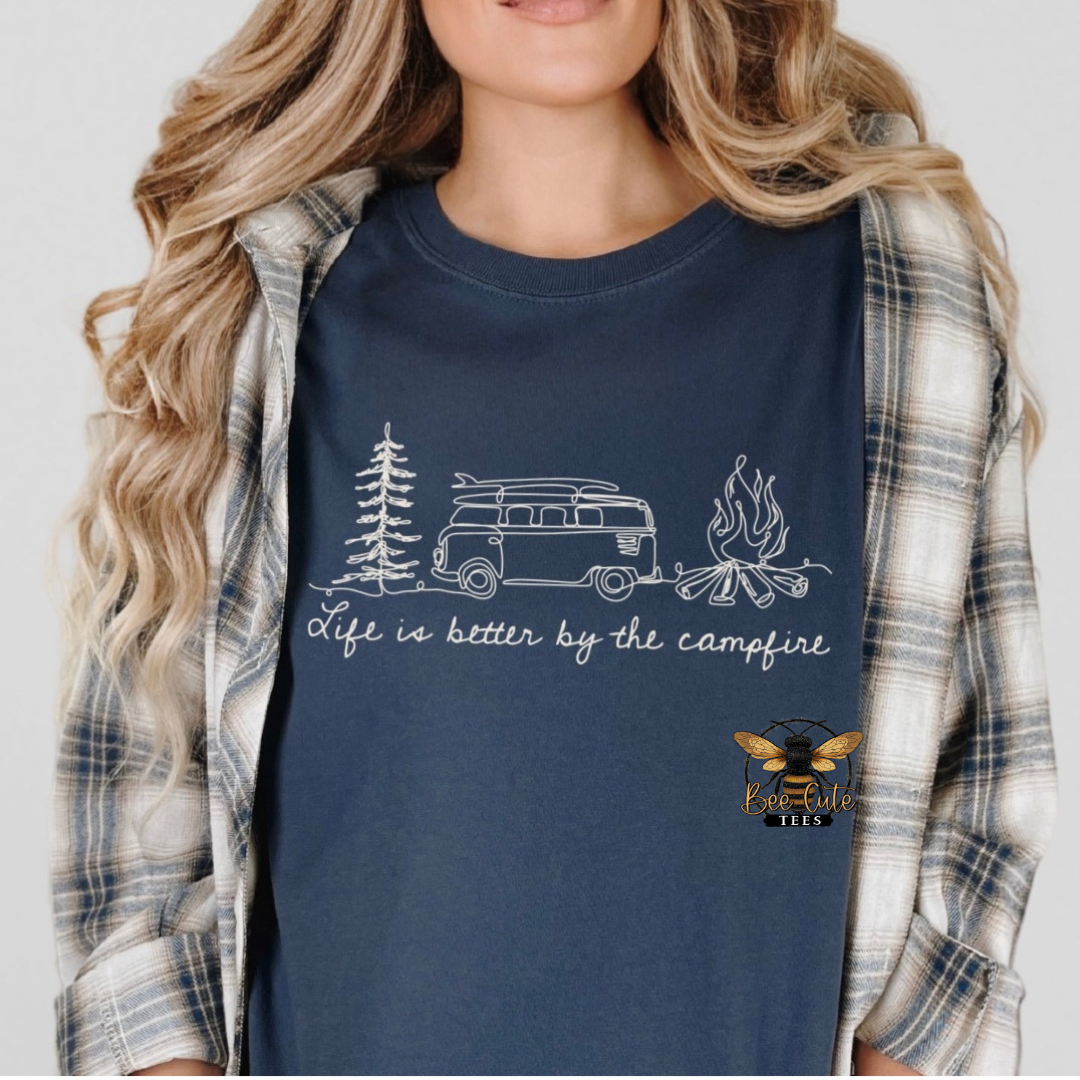 A woman wearing a navy blue graphic t-shirt with a white camping-themed print (van, tree, campfire) and the text 'Life is better by the campfire'. She has a gray and white plaid flannel shirt open over her shoulders.