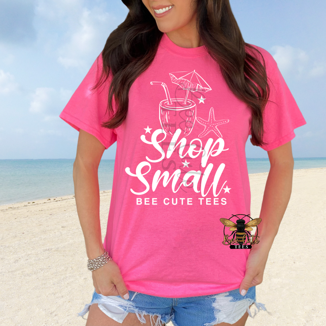 A woman wearing a bright pink graphic t-shirt with white text reading 'Shop Small' and 'BEE CUTE TEES' on a beach background. The t-shirt features a white cocktail graphic with star details.