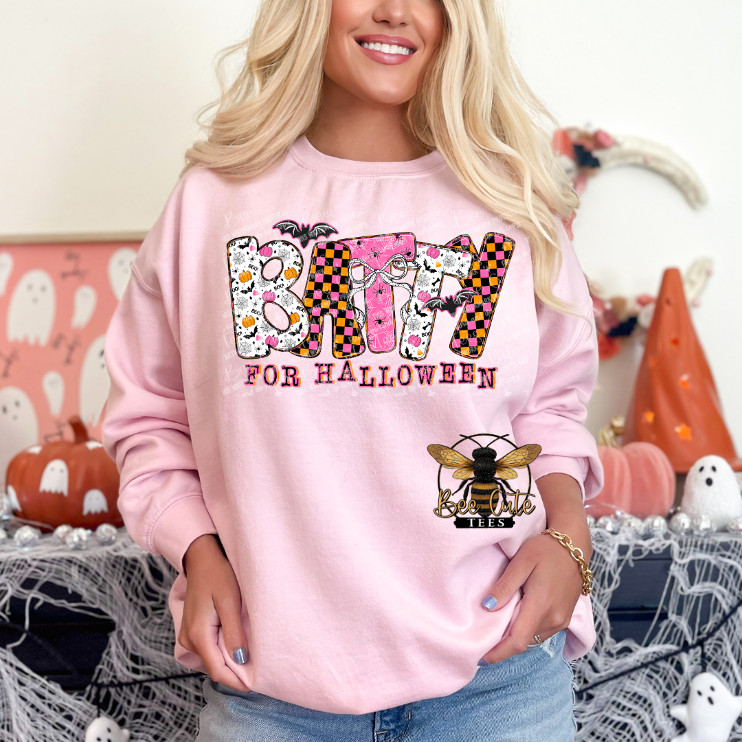 A woman wearing a light pink crewneck sweatshirt with a 'BATTY FOR HALLOWEEN' graphic featuring a bee design, posed in a decorated Halloween room.