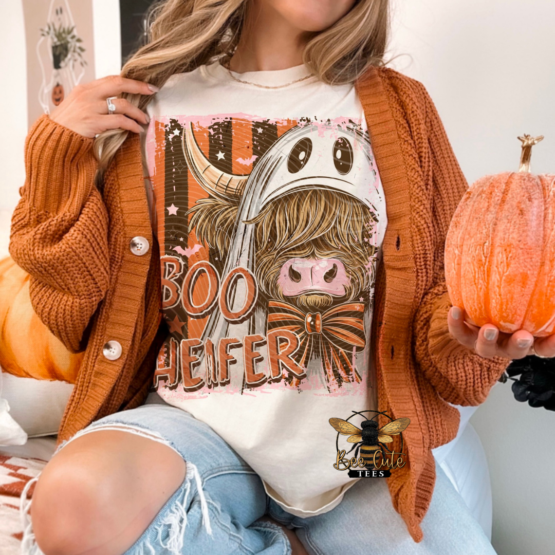 A woman wearing a graphic t-shirt with a cartoon cow in a ghost hat and the word 'BOOHEIFER', styled with an orange knit cardigan and ripped jeans, holding a small orange pumpkin.
