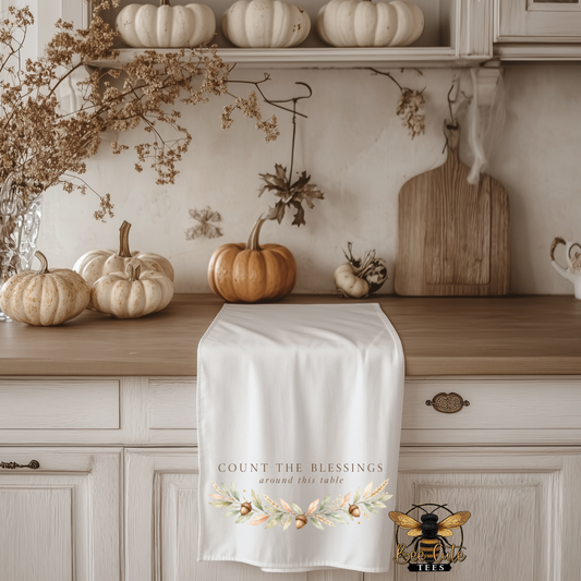 A white kitchen tea towel with the text 'COUNT THE BLESSINGS around this table' and a floral design at the bottom, draped over a wooden kitchen counter decorated with pumpkins and fall decor.