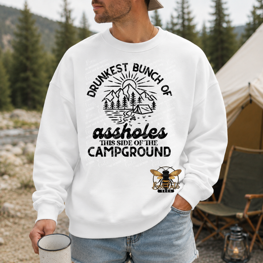 A man wearing a white crewneck sweatshirt with a black graphic that reads 'DRUNKEST BUNCH OF assholes THIS SIDE OF THE CAMPGROUND' and a bee logo, standing outdoors near a tent and camping gear.