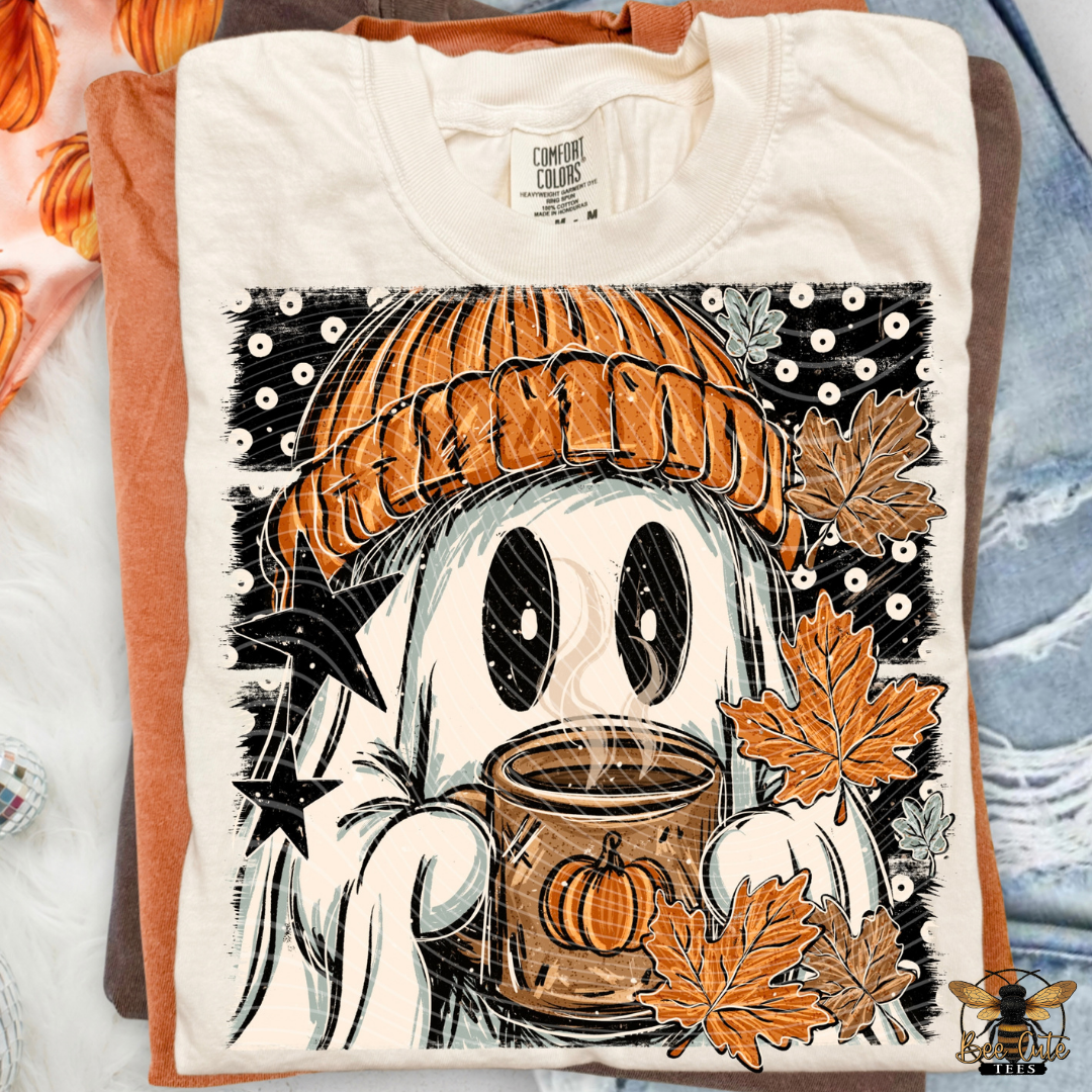A folded cream-colored t-shirt with a brown hem, featuring a graphic print of a cute cartoon ghost wearing an orange knit beanie and holding a pumpkin mug, surrounded by autumn leaves and stars. The shirt is layered over a brown t-shirt and a blue denim jacket on a neutral background.
