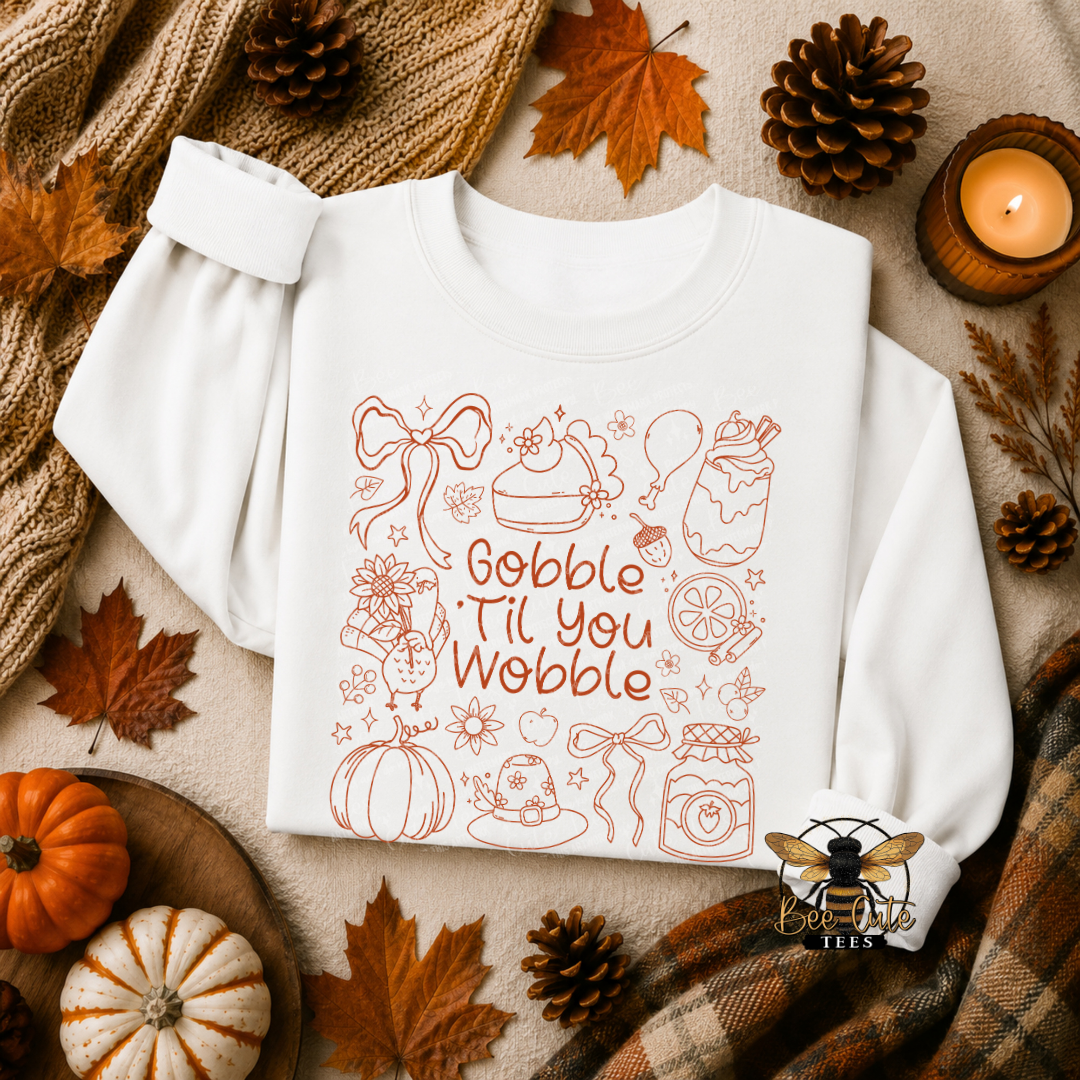 A white long-sleeve sweatshirt laid flat on a neutral surface, decorated with an orange line-art autumn theme and the phrase 'Gobble Til You Wobble.' The sweatshirt is surrounded by seasonal props including pumpkins, pinecones, leaves, a scarf, and a candle.