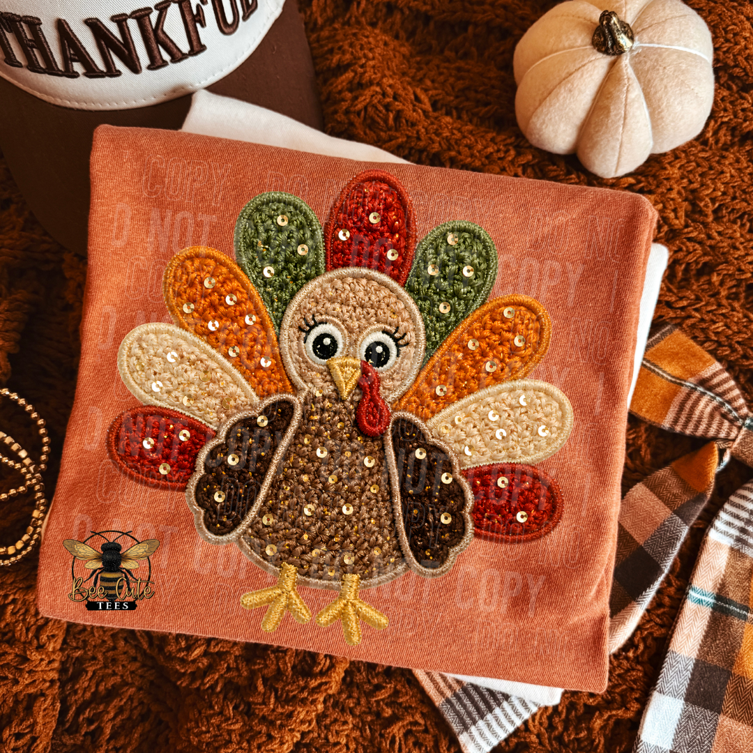 A folded orange t-shirt featuring a faux crocheted and sequined turkey design, styled with autumn props including a small pumpkin, plaid fabric, and a brown knit blanket.