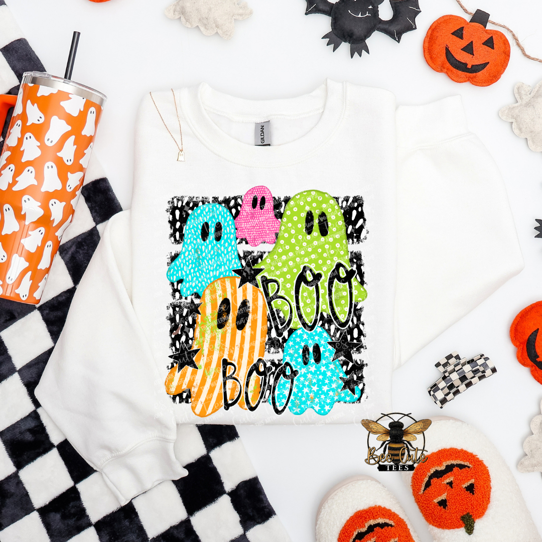 A white crewneck sweatshirt featuring a colorful ' Boo Boo' Halloween graphic with sequined ghosts, laid flat on a white background surrounded by festive props like a ghost cup, jack-o'-lanterns, and black-and-white checkered fabric.