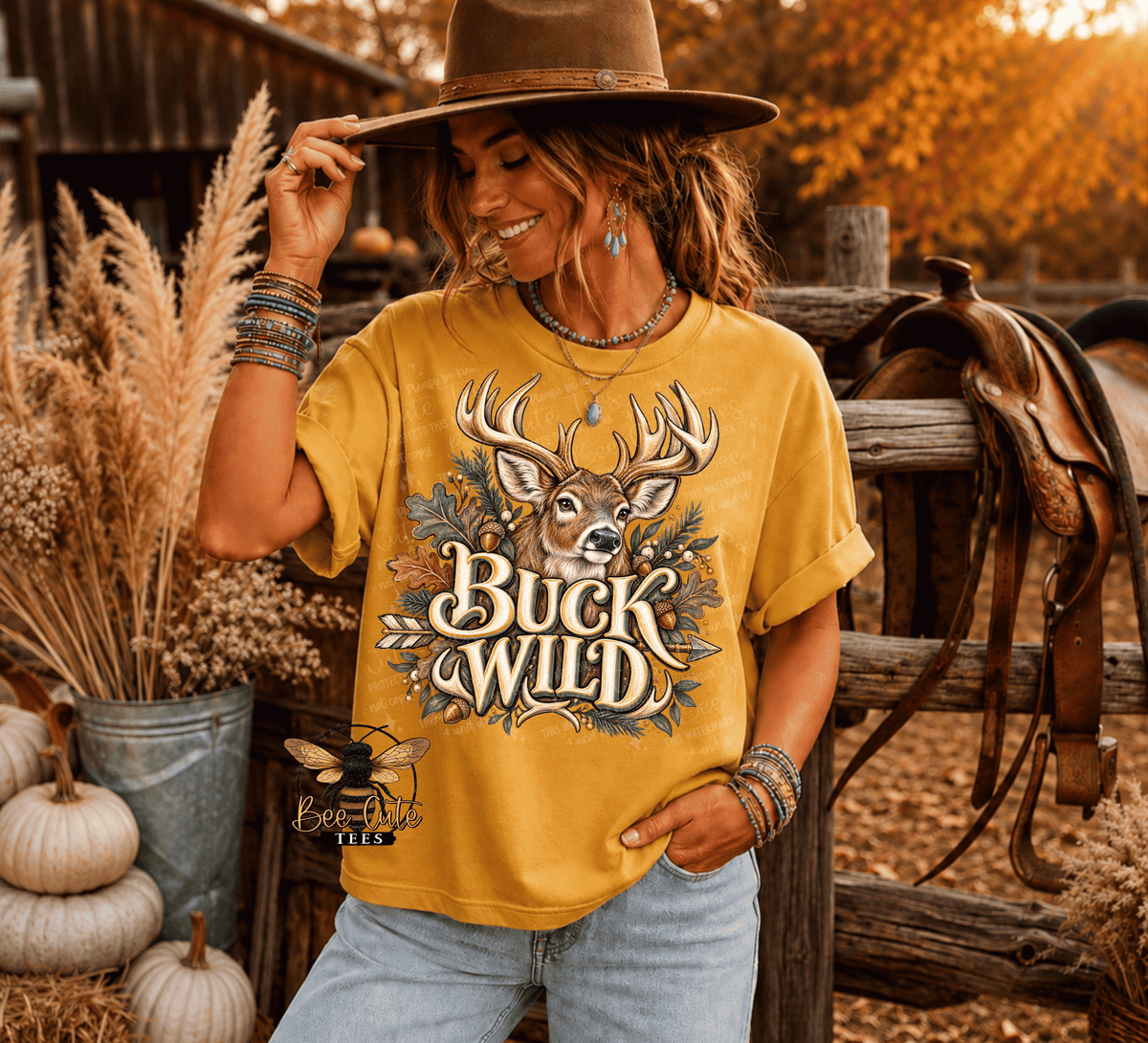 A woman wearing a mustard yellow graphic t-shirt with a deer and the text 'Buck Wild', styled in a rustic autumn outdoor setting with a hat, jeans, and a saddle on a wooden fence.