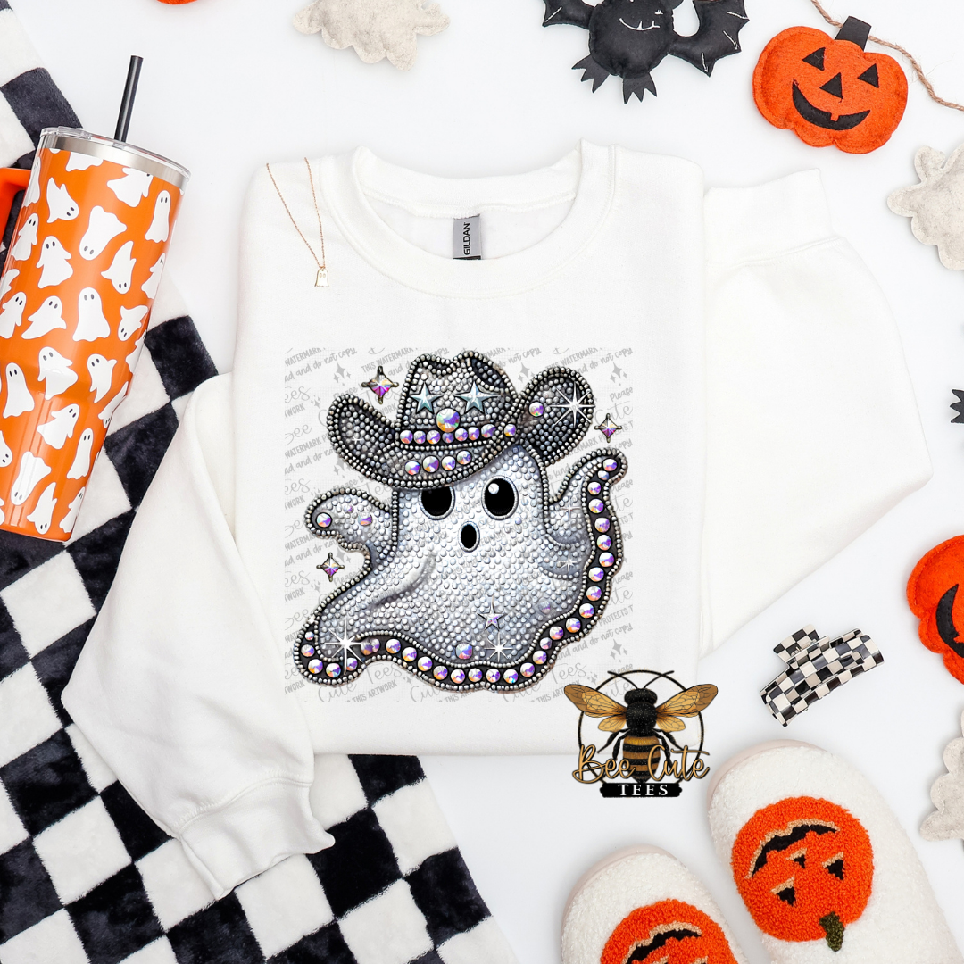 A white long-sleeve sweatshirt laid flat on a white surface, featuring a large glittery ghost graphic wearing a cowboy hat with rhinestone details. The sweatshirt is styled with Halloween props including a black-and-white checkered cloth, an orange cup with ghost illustrations, pumpkins, a bat, a bee ornament, and white slippers.