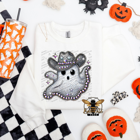 A white long-sleeve sweatshirt laid flat on a white surface, featuring a large glittery ghost graphic wearing a cowboy hat with rhinestone details. The sweatshirt is styled with Halloween props including a black-and-white checkered cloth, an orange cup with ghost illustrations, pumpkins, a bat, a bee ornament, and white slippers.