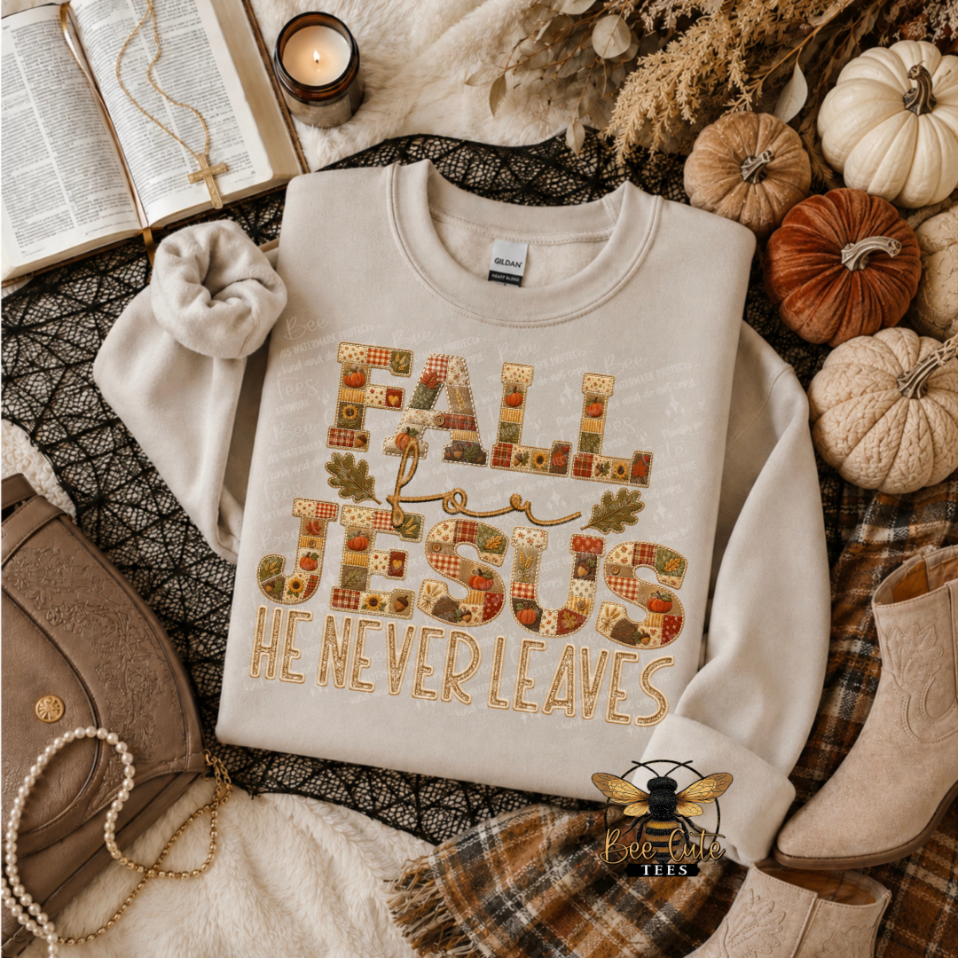 A folded cream sweatshirt with a decorative autumn-themed graphic reading 'FALL FOR JESUS HE NEVER LEAVES' in patchwork-style letters, styled in a cozy flat lay with pumpkins, a plaid scarf, boots, a purse, and an open book.