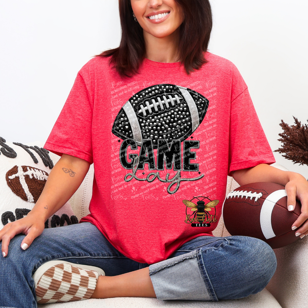 A woman sitting on a couch wearing a heather red graphic t-shirt with a black football design and the words 'GAME Day'. She is holding a brown football and wearing blue jeans.