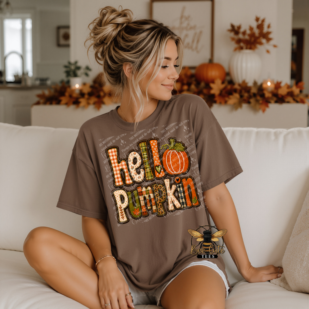 A woman sitting on a white couch wearing a brown graphic t-shirt that says 'hello pumpkin' in a fall-themed patterned font with a pumpkin illustration. The background shows a cozy, decorated living room with autumn leaves and pumpkins.