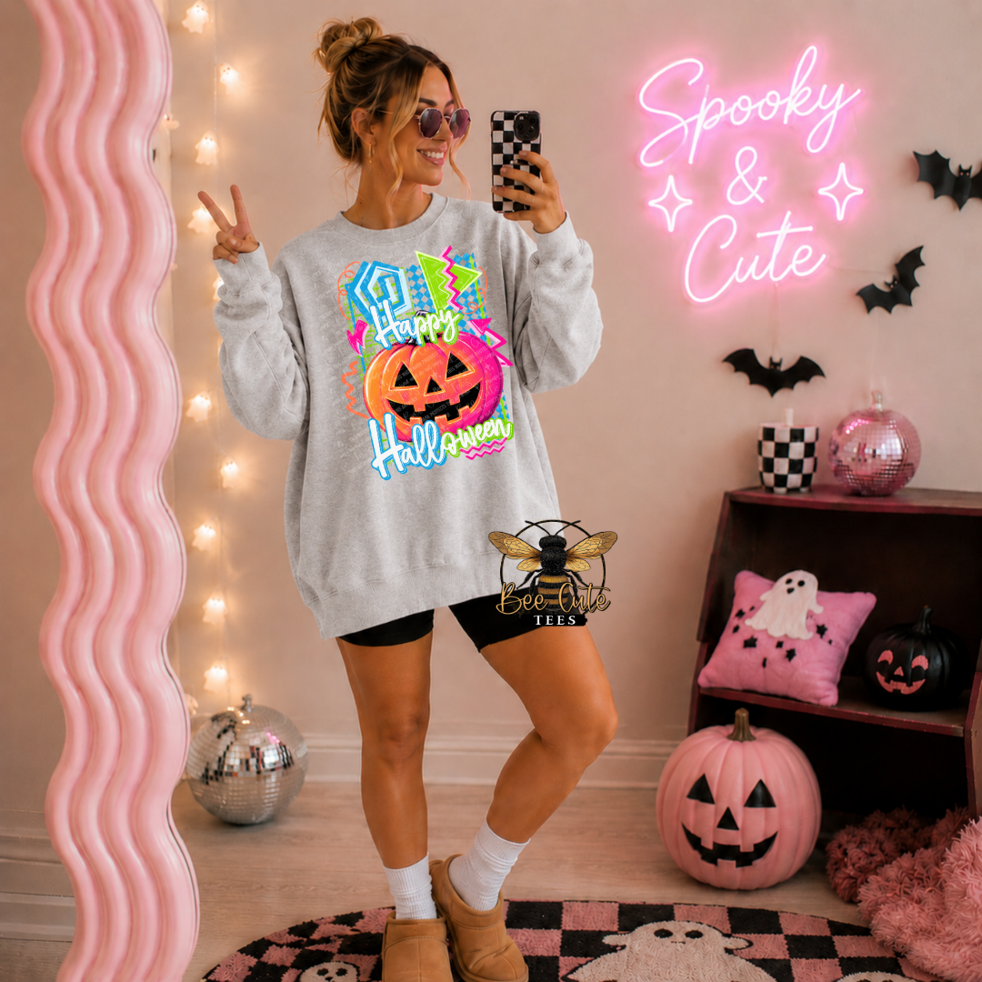 A woman takes a mirror selfie wearing a light gray crewneck sweatshirt with a colorful 'Happy Halloween' graphic featuring a jack-o'-lantern. The room is decorated for Halloween with a pink wavy mirror, a pink neon sign that reads 'Spooky & Cute', pumpkins, and black-and-white checkered accents.
