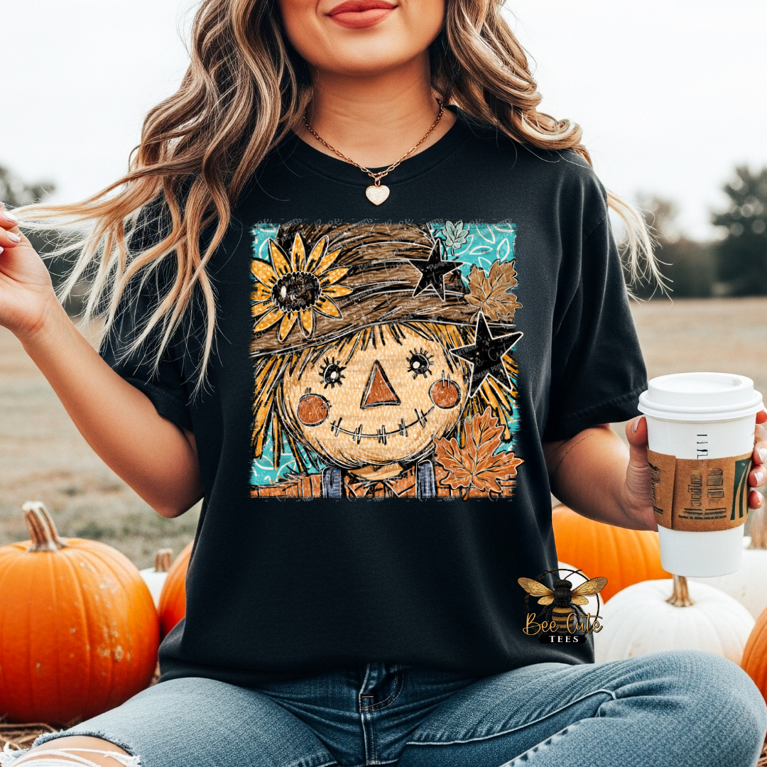 A woman wearing a black graphic t-shirt with a colorful autumn scarecrow design, sitting in a pumpkin patch and holding a coffee cup.