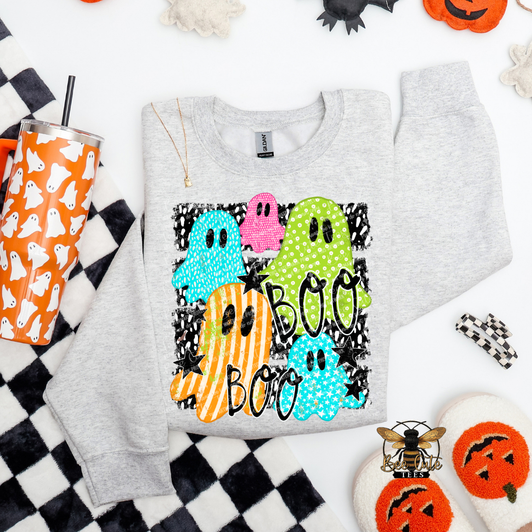 A light heather gray crewneck sweatshirt with a colorful Halloween graphic featuring four ghosts and the word 'BOO BOO' in black script. The sweatshirt is styled in a flat lay with autumn-themed props including a checkered blanket, a tumbler, and pumpkin decorations.