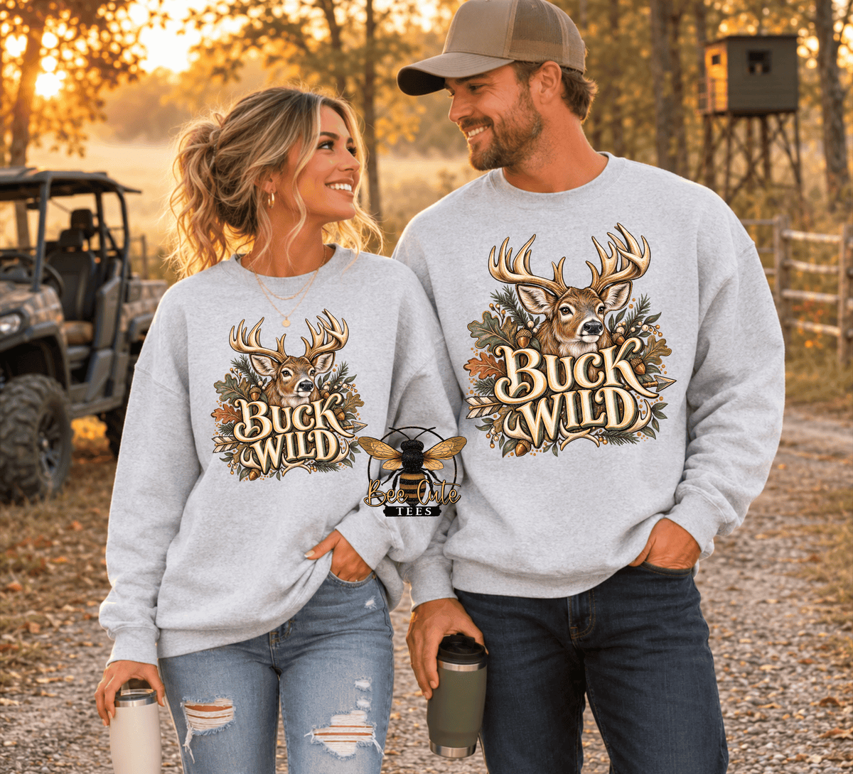 A man and a woman standing outdoors in a wooded setting at sunset, both wearing matching light gray crewneck sweatshirts with a large deer graphic and the words “Buck Wild” on the front. The woman also wears ripped blue jeans, and the man wears dark jeans and a tan cap.