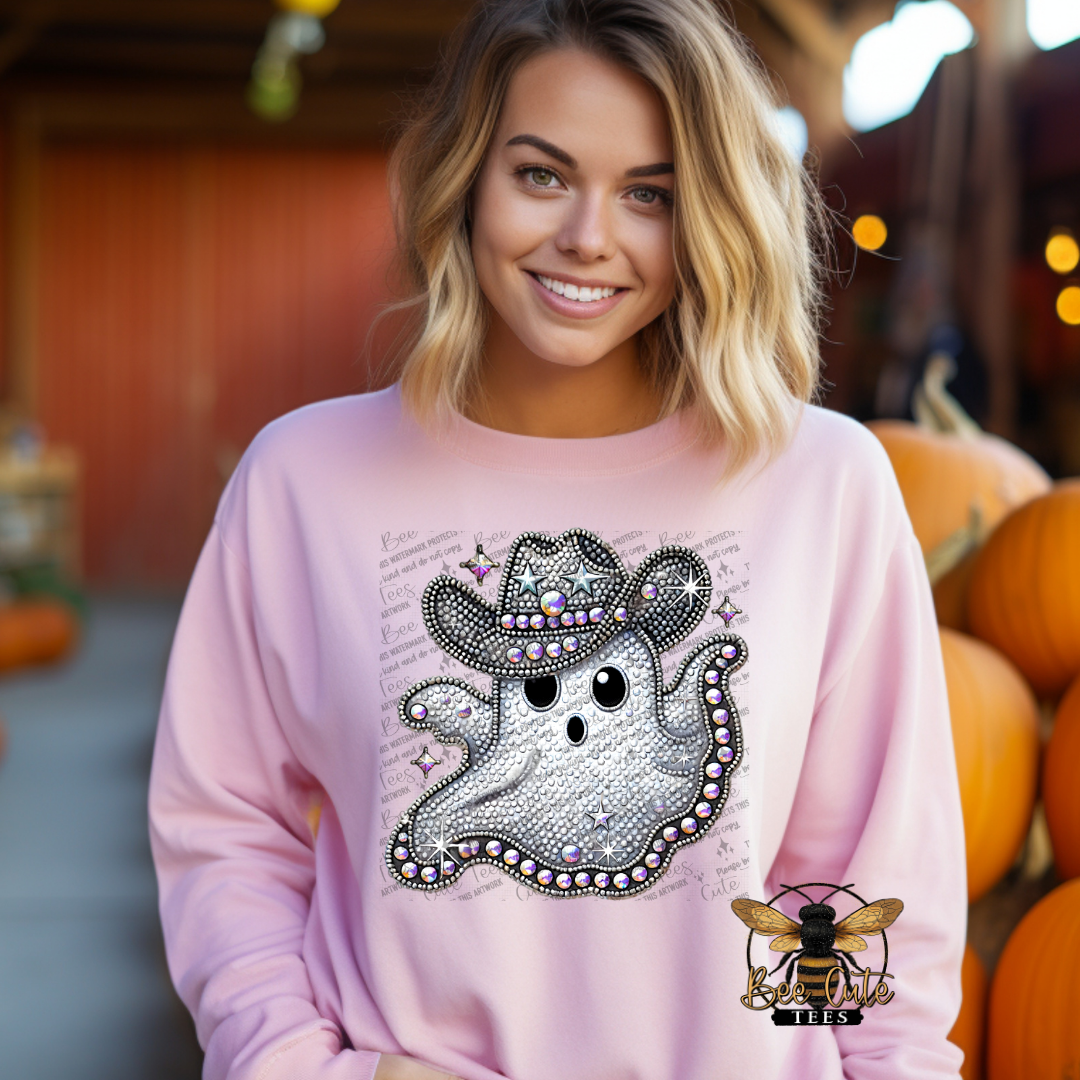 A smiling blonde woman wears a light pink long-sleeve sweatshirt with a large glittery silver ghost graphic on the front, posed in front of a rustic barn-like setting with pumpkins.
